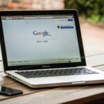 A MacBook Pro displaying Google Search on a wooden table outdoors, next to a smartphone.